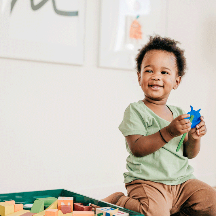 Joyful toddler sitting on the floor with a toy, showcasing the playful aspect of speech therapy for toddlers.